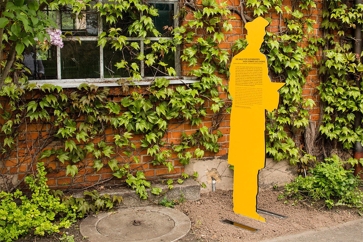 A yellow cut out figurine standing infront of the narrow-gauge railway hall overgrown with ivy
