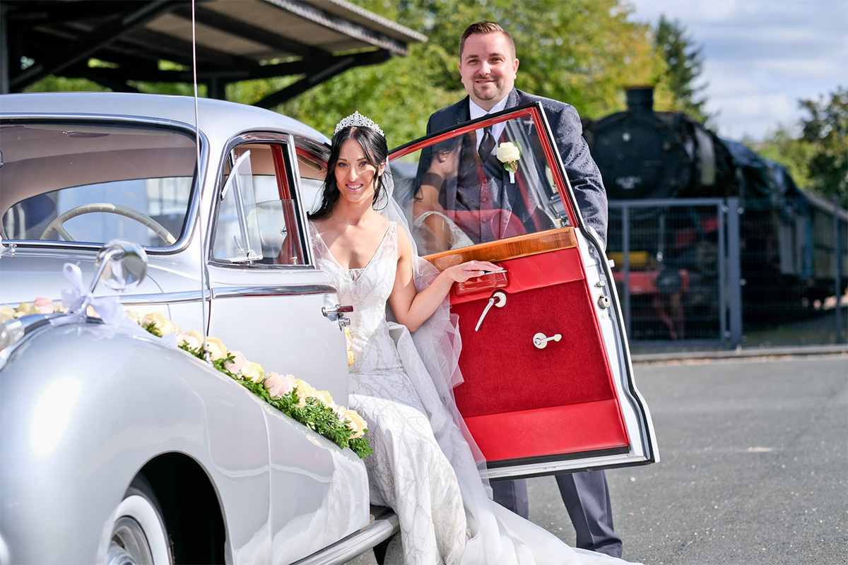 Der Bräutigam öffnet die Tür eines silbernen Oldtimers für die Braut